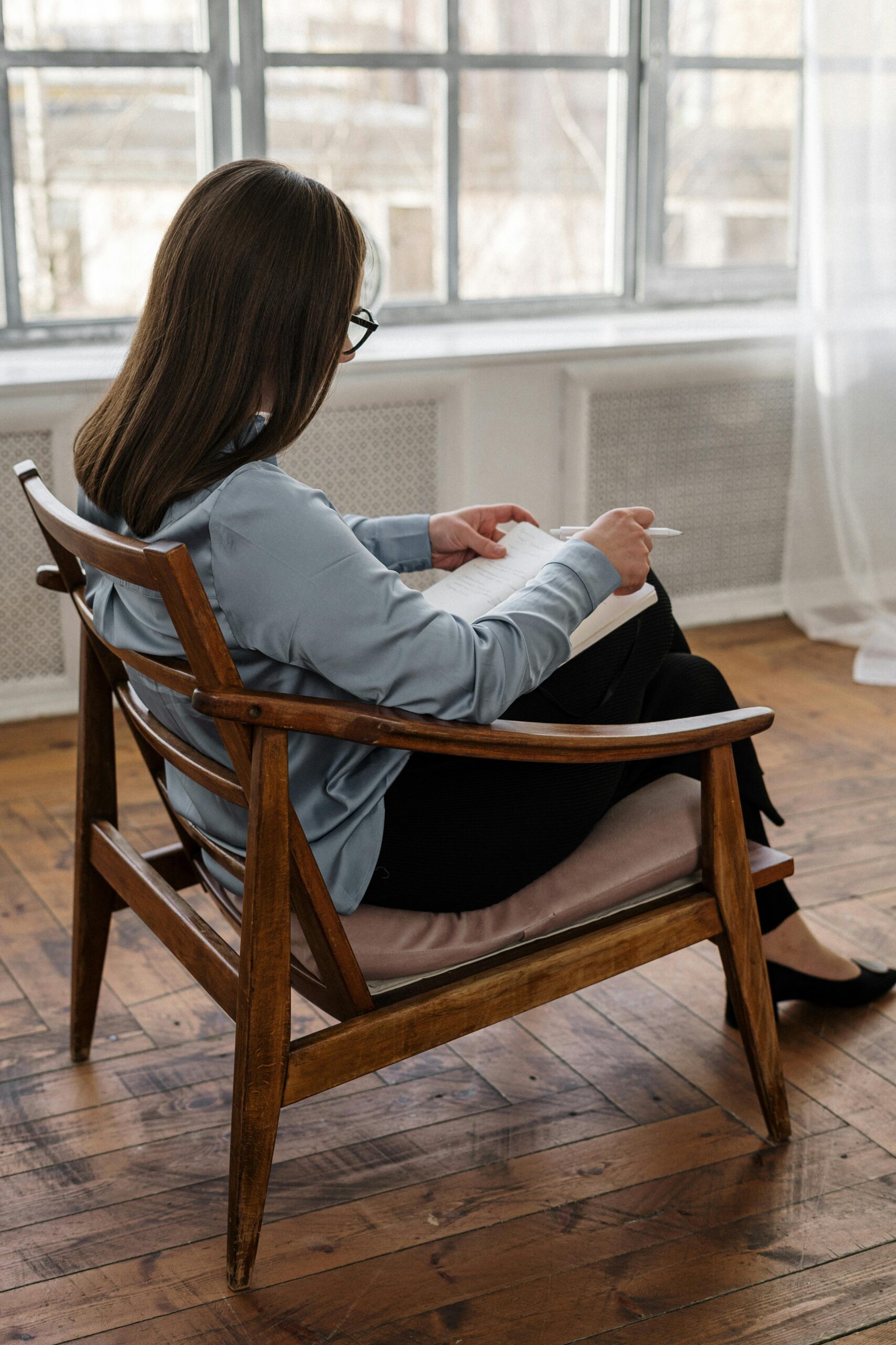 Woman with glasses sitting in a wooden chair while writing in a notebook, capturing thoughtful moments.